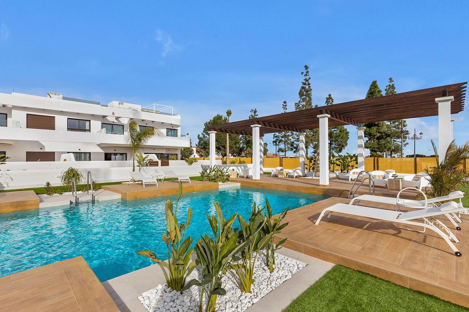 Resort-style pool area with white buildings, a wooden deck, and a large pergola under a bright blue sky.