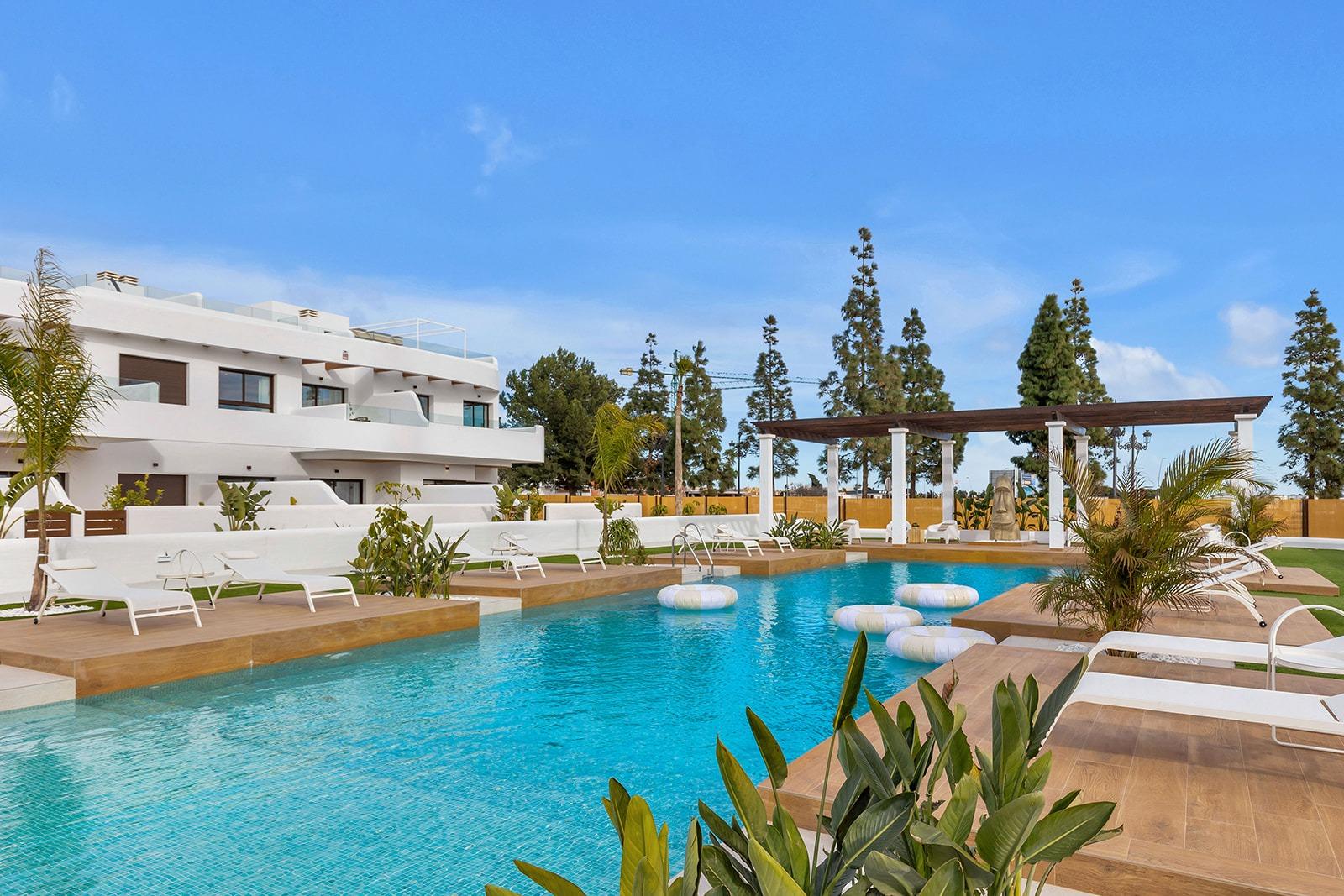 Resort pool area with white modern buildings, wooden decks, white lounge chairs, and palm plants under a blue sky.