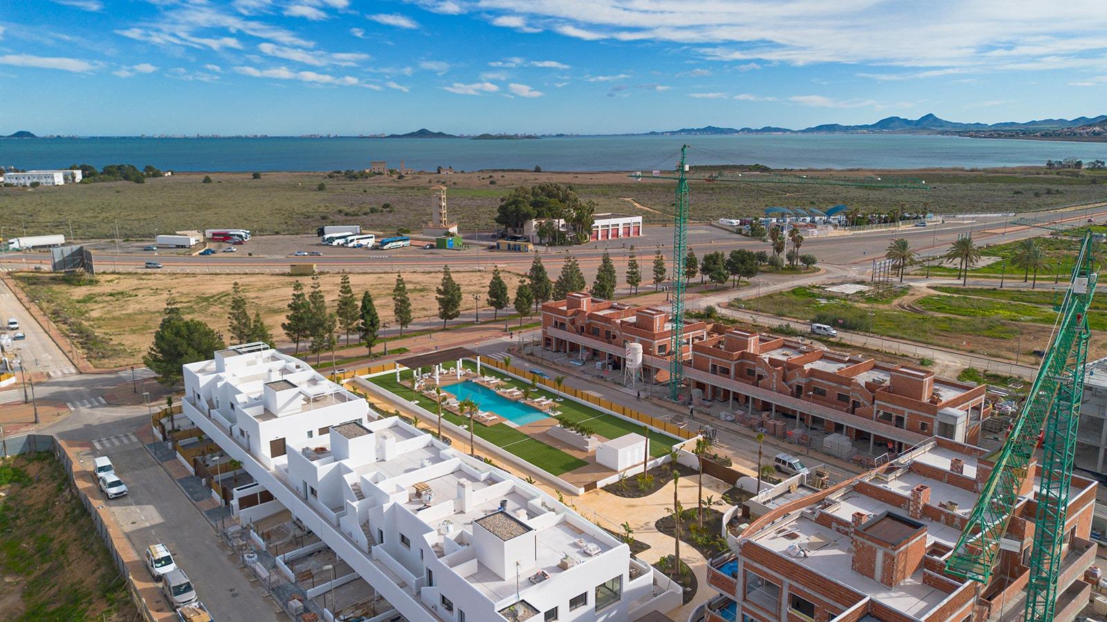 Aerial view of a coastal construction site with white modern buildings, a pool, brick buildings under construction, cranes, and a road with trucks and cars; sea in the distance.