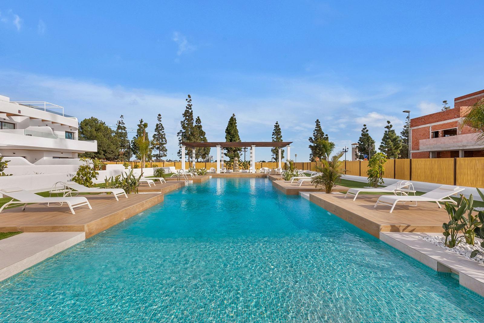 Crystal blue rectangular pool surrounded by white lounge chairs and palm trees in a sunny outdoor resort setting.