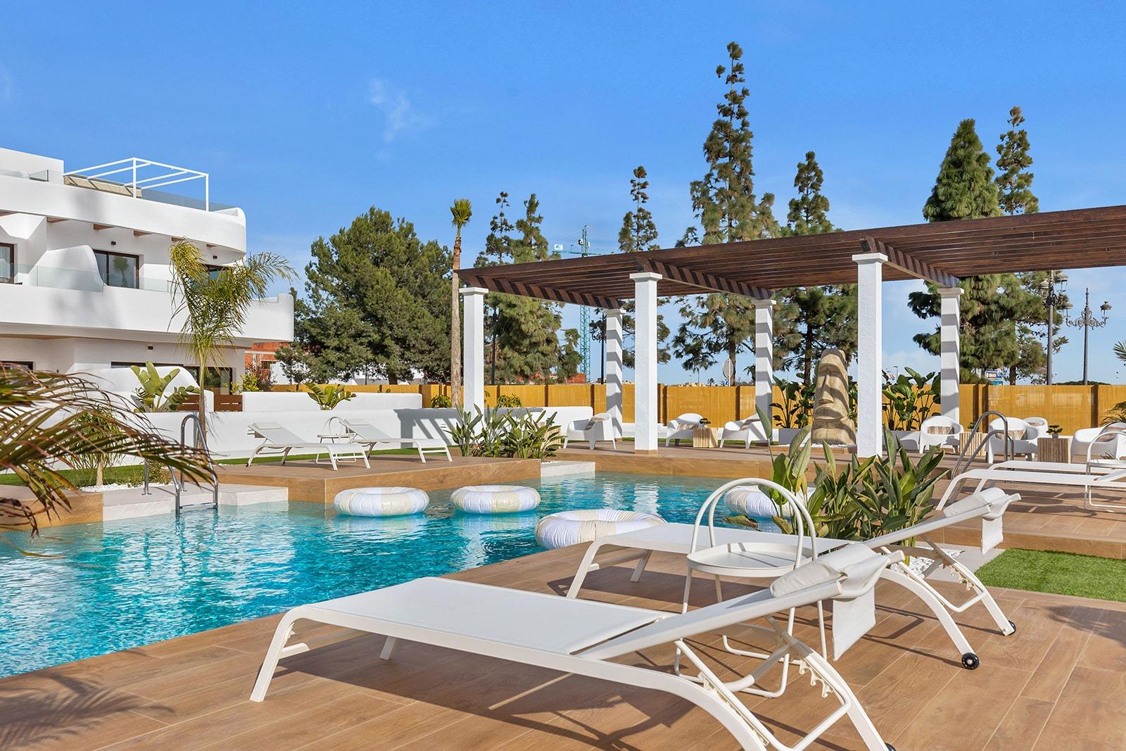 Poolside resort scene with white loungers, inflatable rings, and a wooden deck under a pergola against a blue sky
