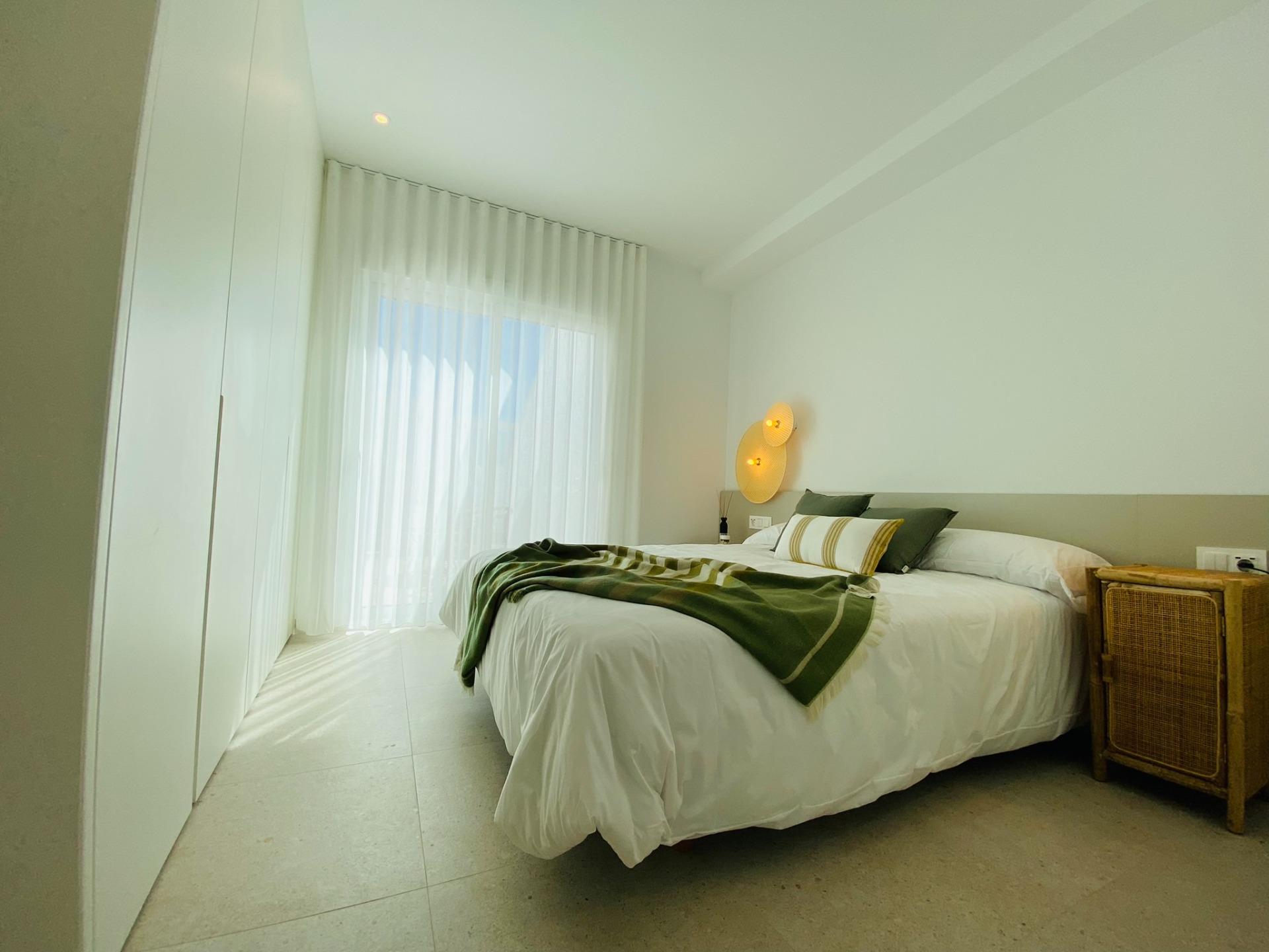 Bright modern bedroom with a large bed, white linens, and a green blanket and pillows by a wicker nightstand with a sheer-curtain window nearby