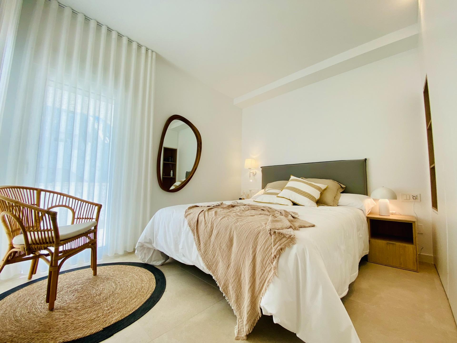 Bedroom with a made bed, beige throw, striped pillows, and a round wooden-framed mirror on the wall opposite a sheer-curtain window with wicker chairs nearby.