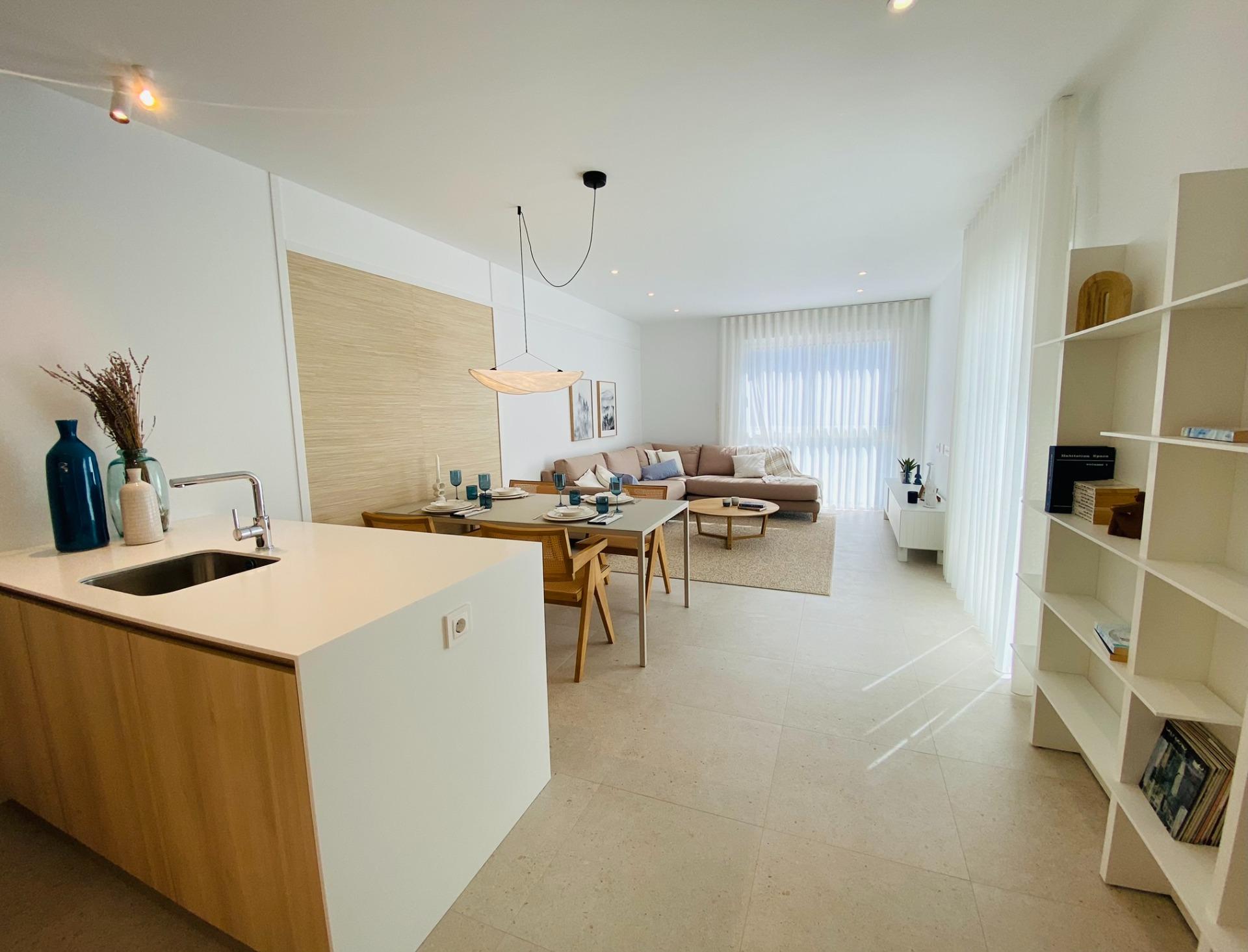 Bright, airy open-plan living area with a white kitchen island, wooden cabinets, and a dining table set near a beige sofa and large windows with sheer blinds in the background.