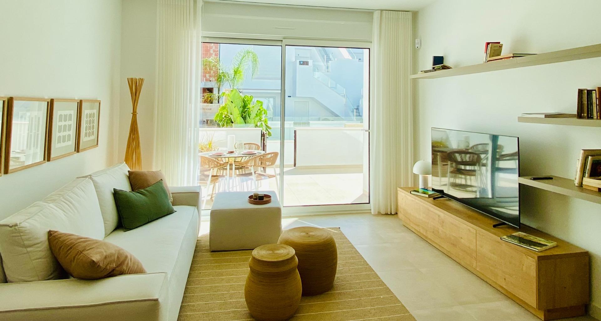 Bright modern living room with white sofa and beige cushions, facing a wooden TV stand and glass doors to a sunlit balcony.