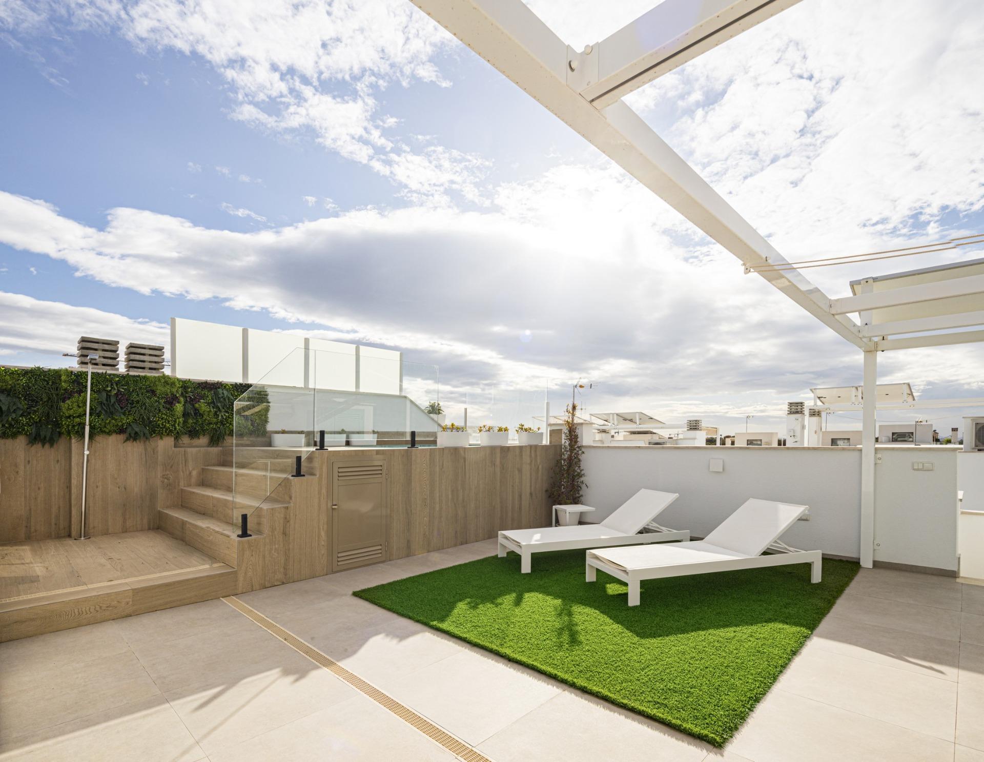 Rooftop terrace with white lounge chairs on a small green grass patch, wooden steps, and glass railing under a blue sky.
