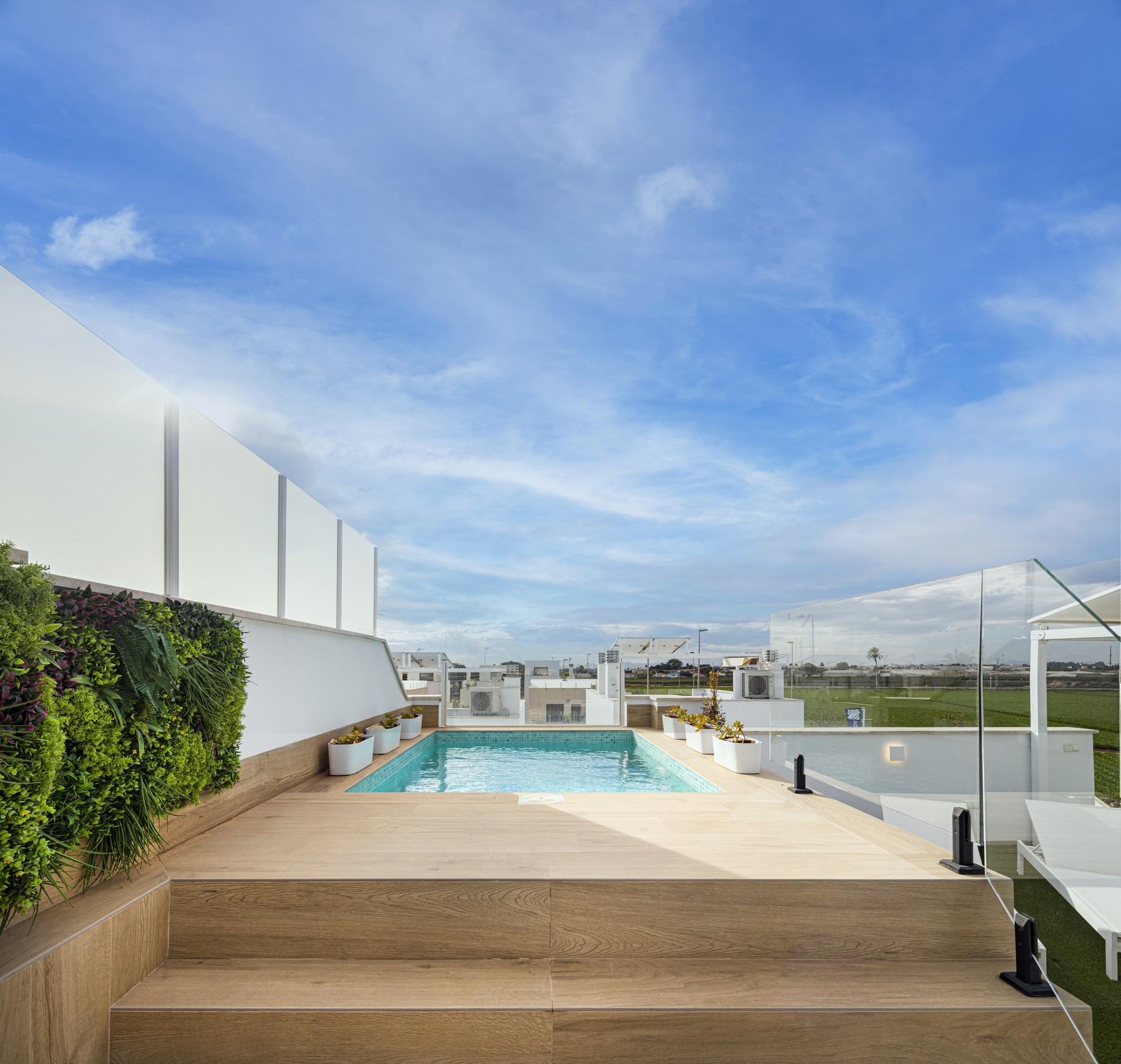 Modern rooftop patio with a rectangular pool, wooden deck, glass railing, and lush green plants along the left wall under a blue sky.