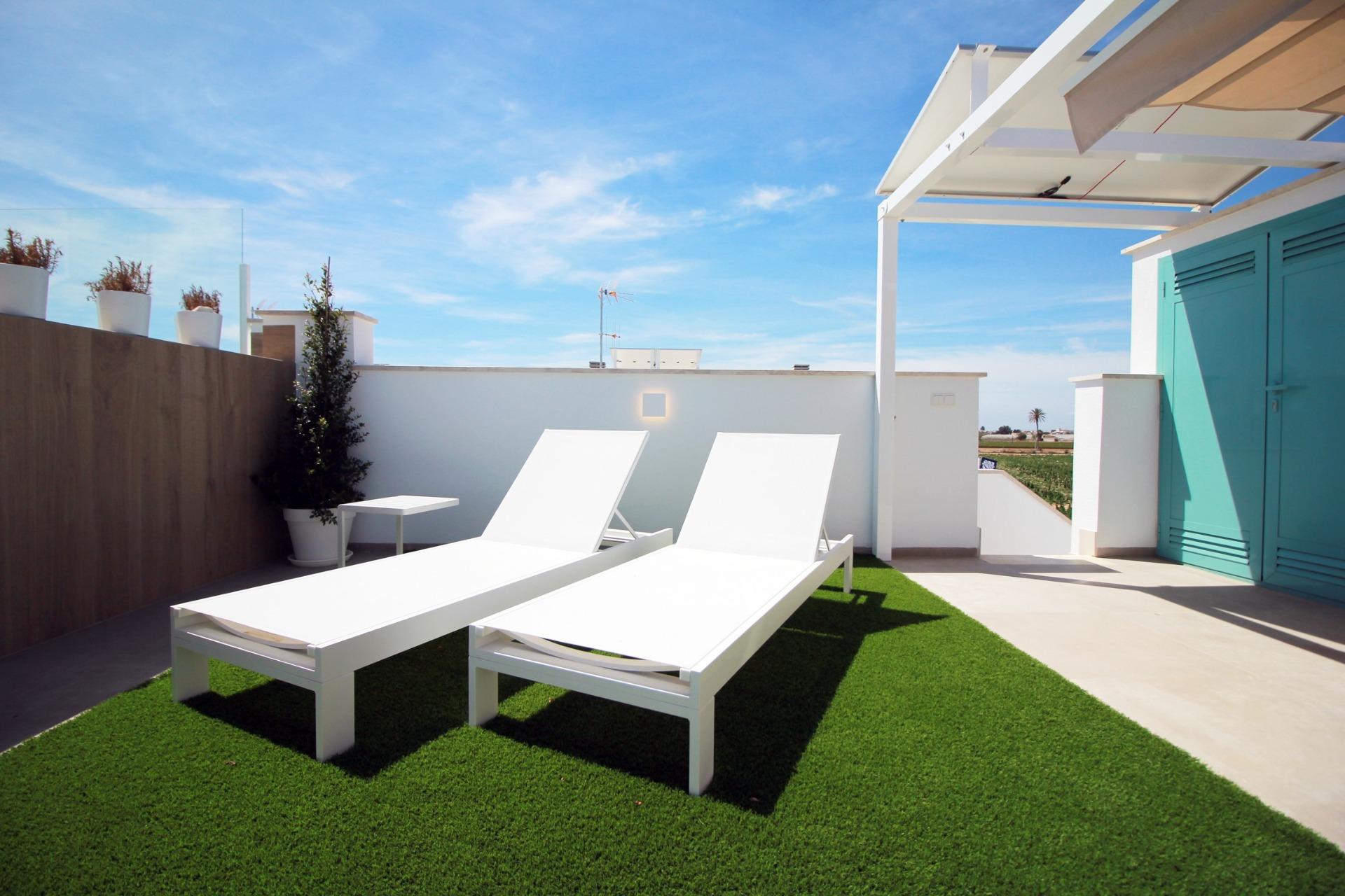 Rooftop patio with two white lounge chairs on green artificial grass under a bright blue sky; turquoise door on the right side.