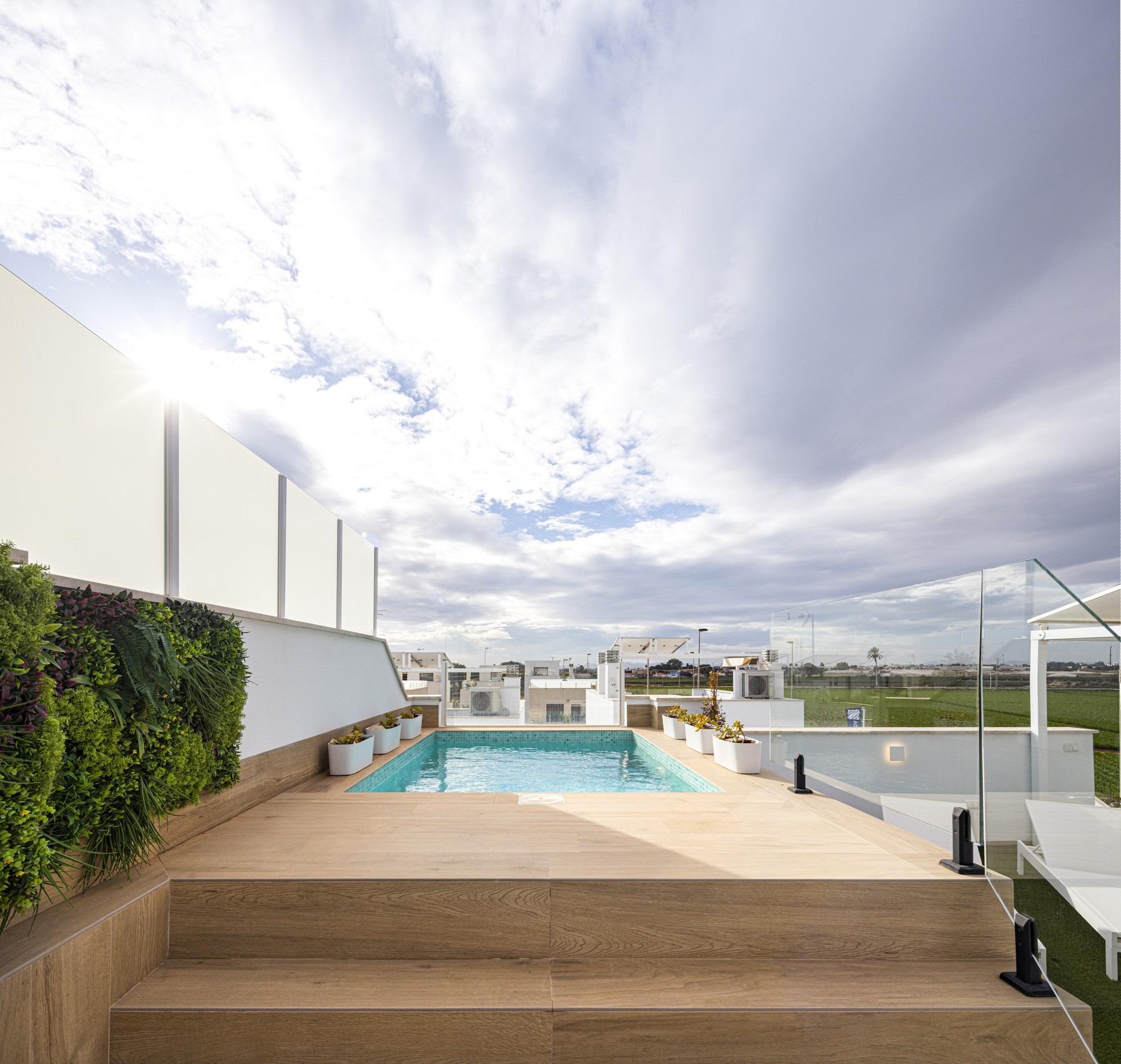 Rooftop deck with a rectangular pool, wooden steps, glass railing, and green plants against a cloudy sky