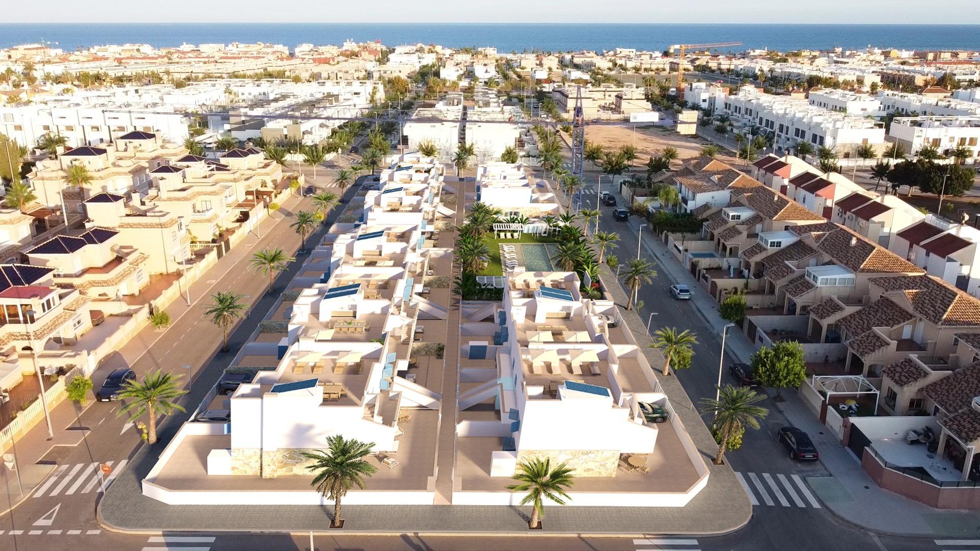 Aerial view of a coastal residential complex with white modern houses, palm trees, and a central pool courtyard near the sea.