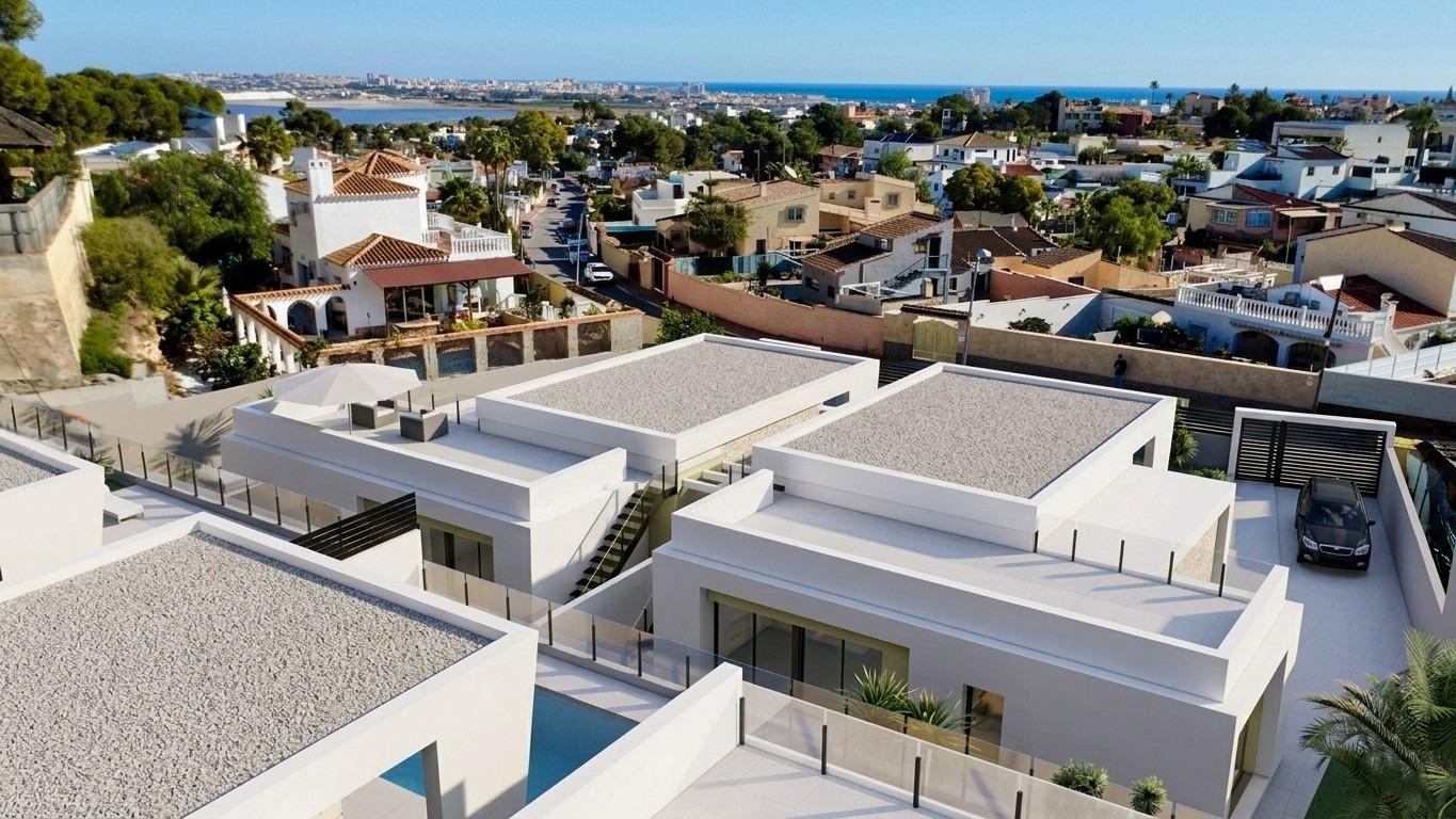 Aerial view of white modern villas with flat roofs and glass railings, near a coastal town with the sea in the background