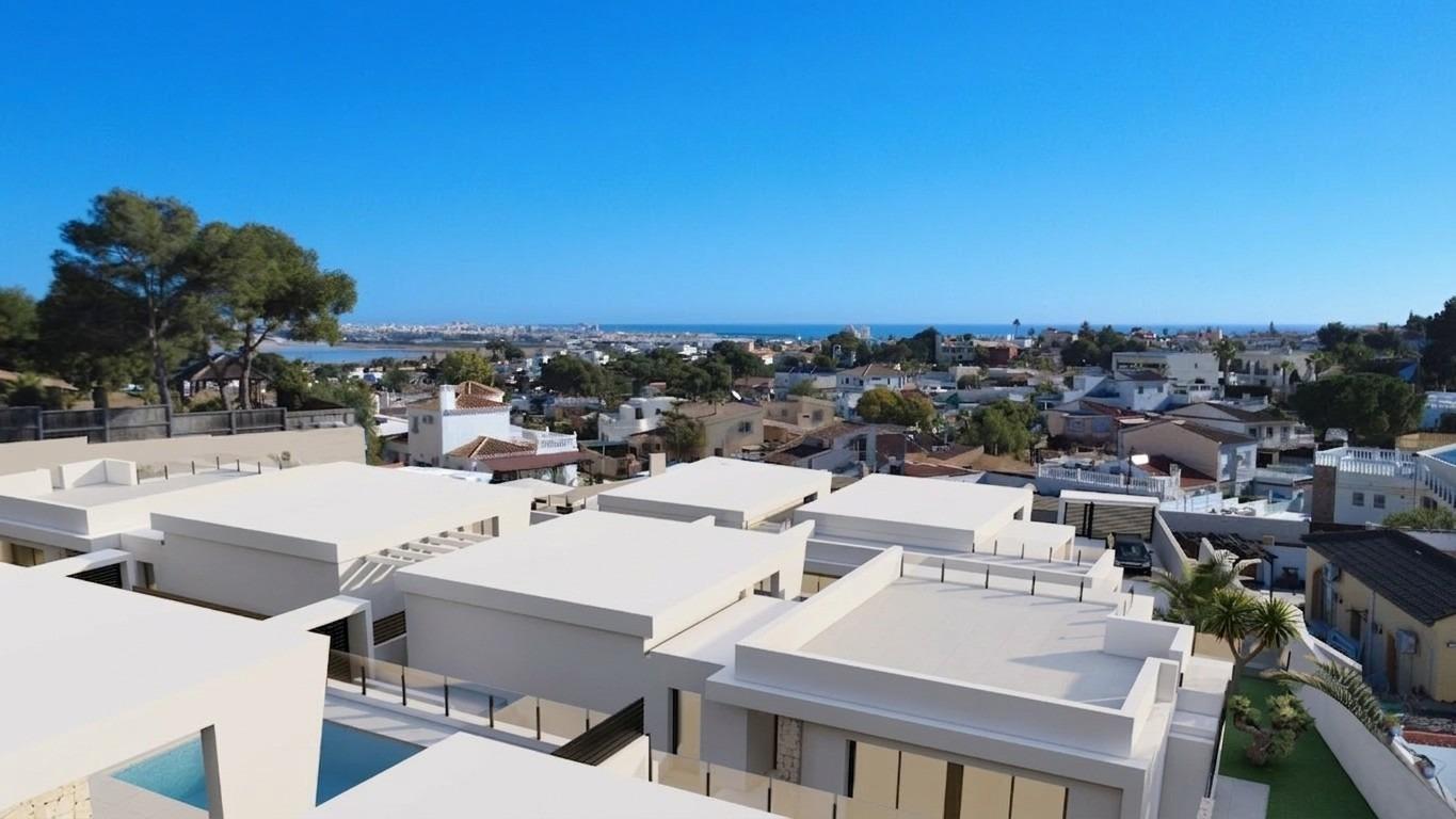 Aerial view of white flat-roofed houses clustered in a coastal town, with the sea on the horizon under a blue sky.