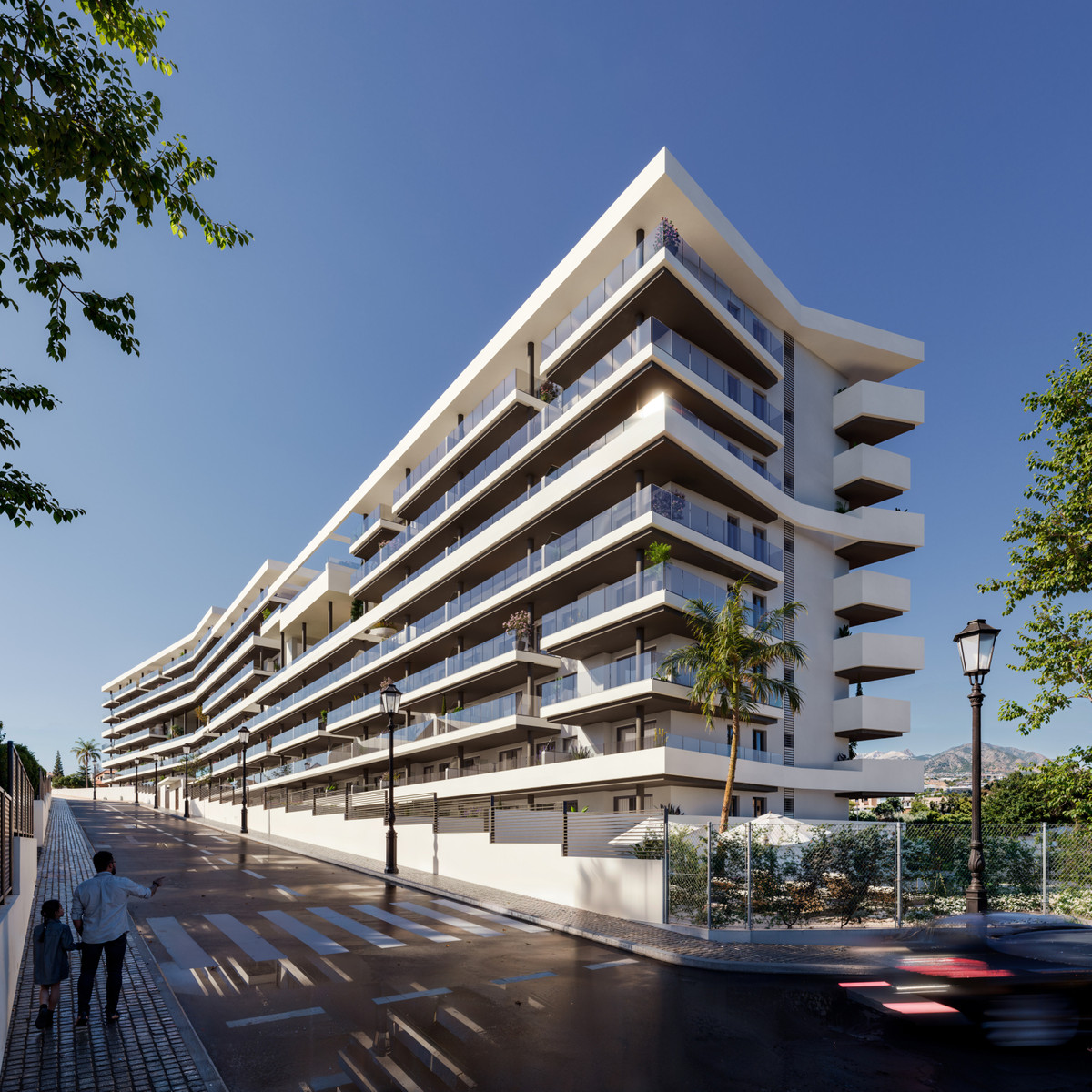 Modern white apartment building with tiered balconies under a clear blue sky; palm trees and street lamps along a pedestrian crossing nearby.
