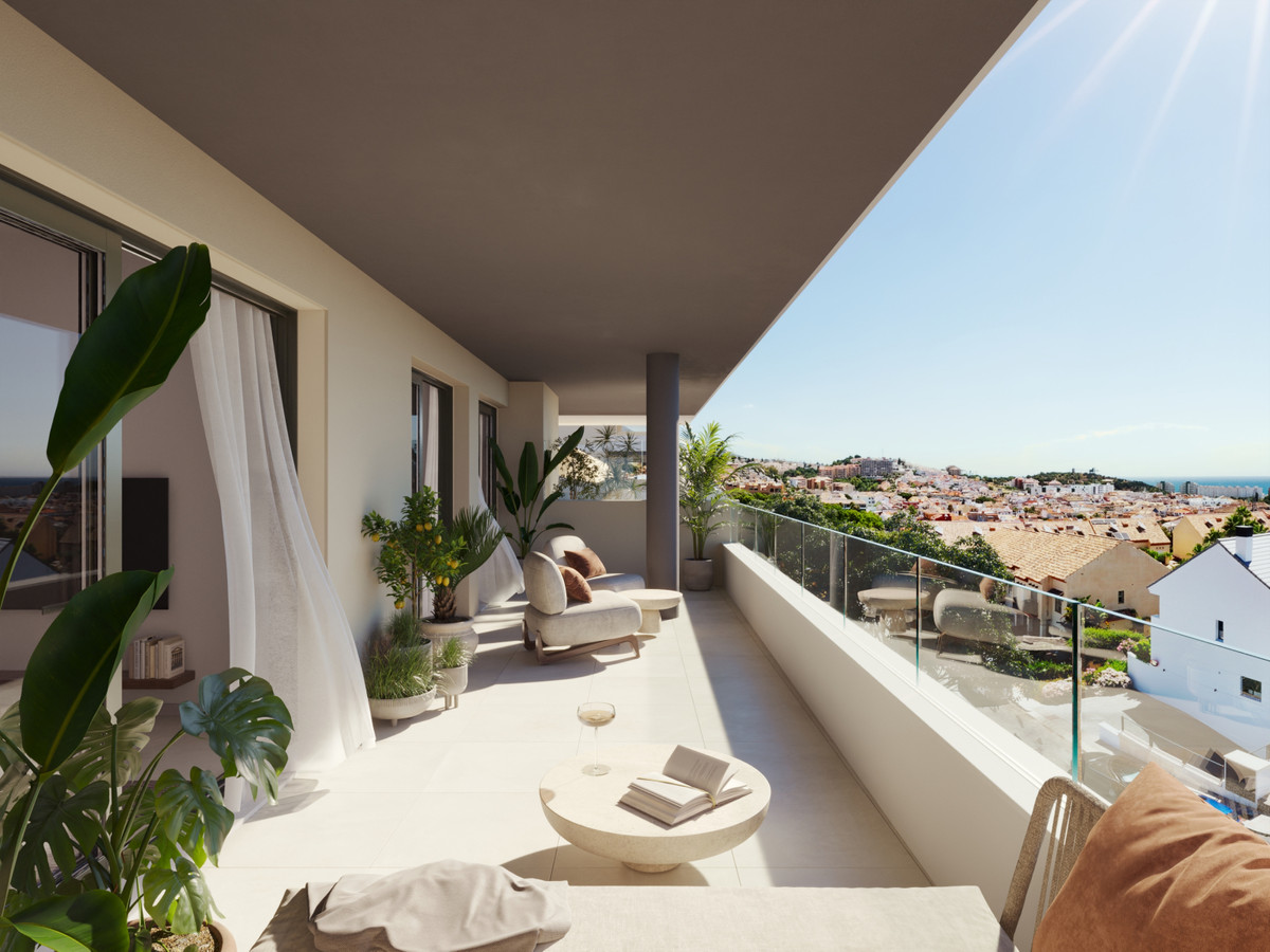 Sunny apartment balcony with white tiles, potted plants, and a cozy seating area overlooking a coastal town.