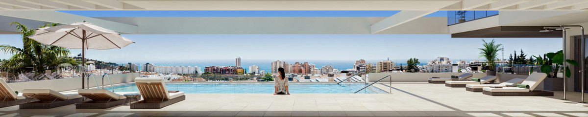 Rooftop pool overlooking a coastal city, with lounge chairs, umbrellas, and a person sitting at the pool edge.